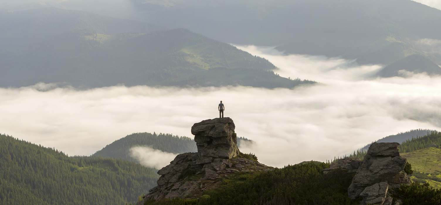 Un homme contemplant l'immensité depuis un sommet rocheux, entouré de collines verdoyantes et de nuages embrassant l'horizon.
